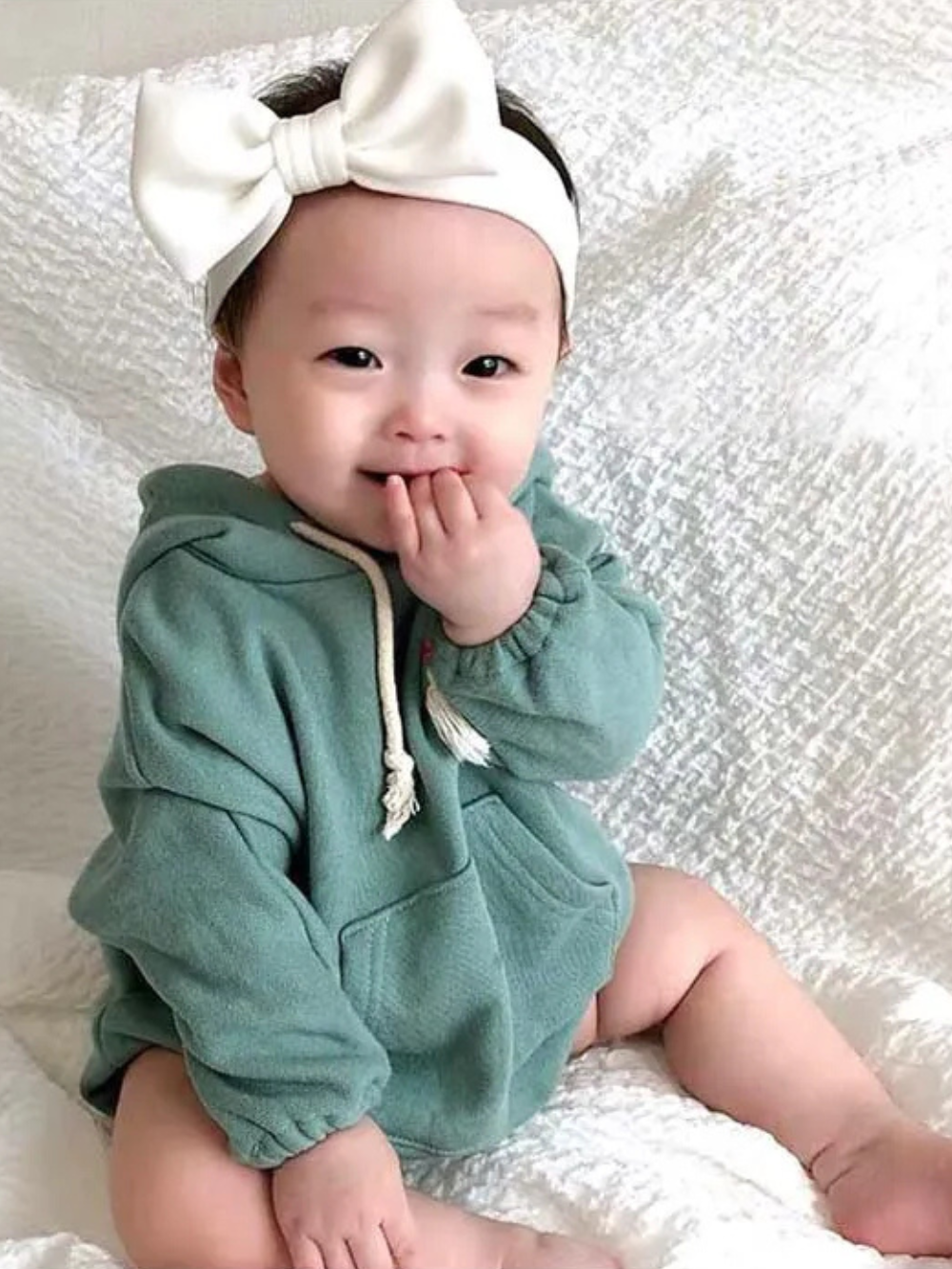 大きな白いリボンヘアバンドを付けてグリーンのフード付きコットンボディスーツを着た赤ちゃんが座っている写真
