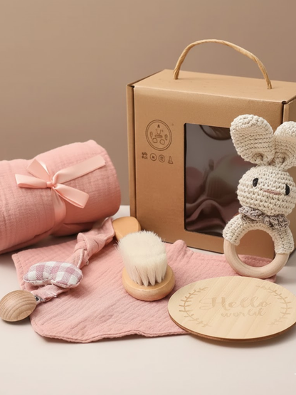 ピンク系のベビーギフトセット。うさぎ型のかぎ針編み歯固め、柔らかいブラシ、綿の布、木製プレートがクラフトボックスの前に並べられている。