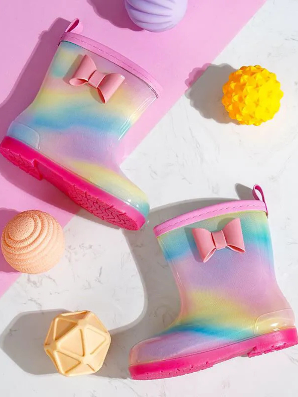 ピンクソールのグラデーションレインボーベビーレインブーツをおもちゃと一緒に並べたトップビュー写真
