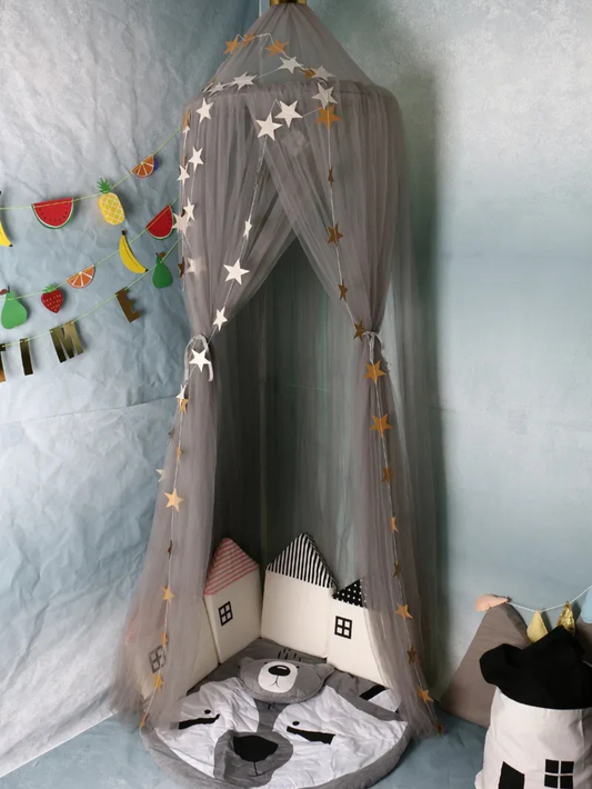 グレーのメッシュ天蓋に星型ガーランドを飾り、床にクッションとラグを敷いたコーナープレイスペースのイメージ（星飾り付きメッシュキャノピー）

