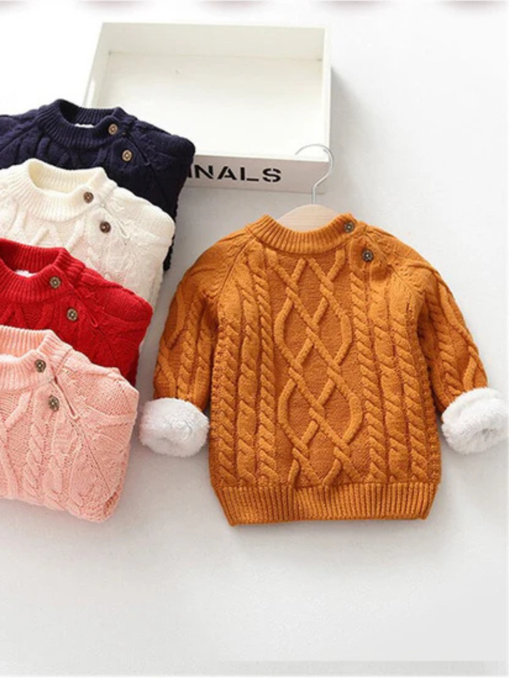 オレンジの裏ボア付きケーブル編み子どもニットセーターと畳まれたカラーバリエーションのフラットレイ
