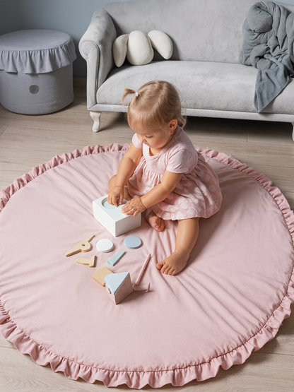 リビングの床に敷いたピンクの円形フリル付きベビープレイマットの上で座って遊ぶ幼児 木製おもちゃと一緒の使用イメージ
