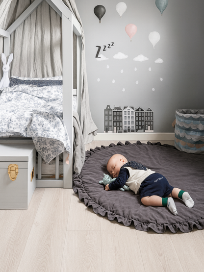 子ども部屋の床に敷いたダークグレーの円形フリル付きベビープレイマットの上で寝転ぶ赤ちゃん 室内の使用イメージ
