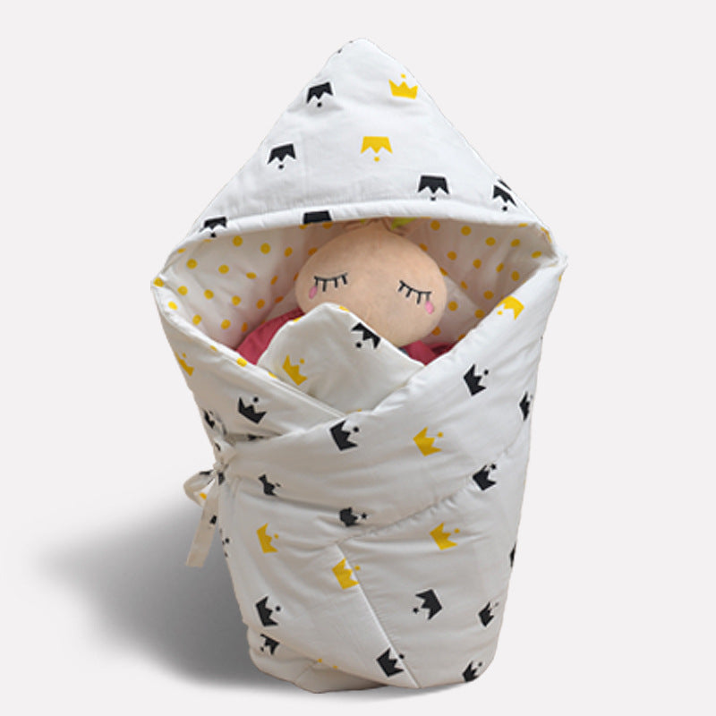 ホワイト地にイエローとブラックの王冠柄が入ったベビーおくるみにぬいぐるみの赤ちゃんがくるまれている写真（ベビーおくるみ 王冠柄 ホワイト）
