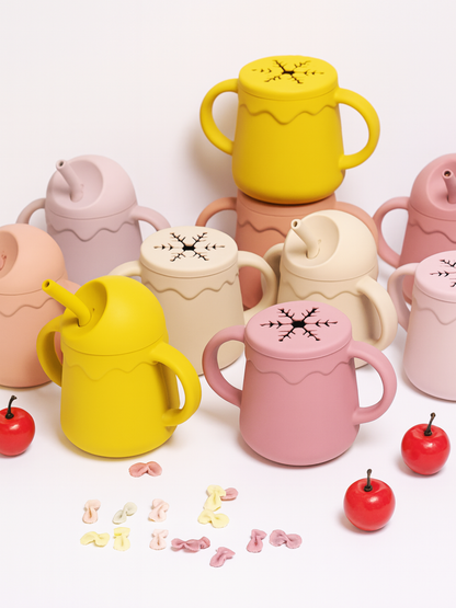 ピンクとイエローのシリコン2in1ノースピルベビーカップが並び、前に小さなお菓子とリンゴ型のおもちゃが置かれている画像（シリコンベビーカップ）
