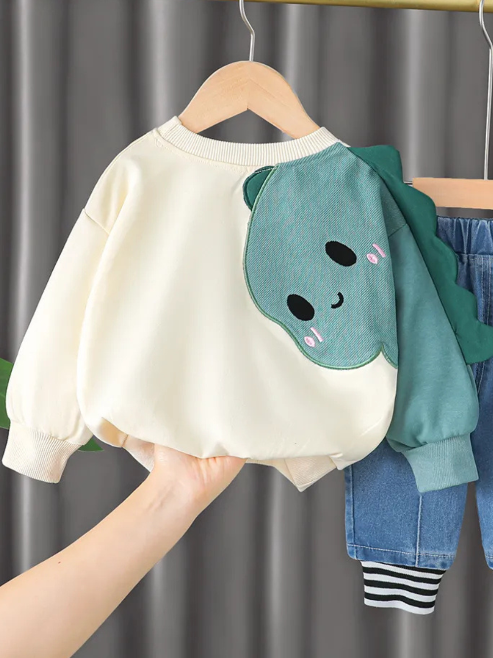 恐竜スリーブ付きクリームカラー長袖トレーナーをハンガーに掛けて後ろ姿を手で支えている画像
