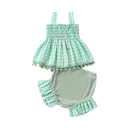 白背景のスタジオ撮影で、グリーンのギンガムチェック柄スモッキングトップスと、セージグリーンのリブ素材ショートパンツのベビー2点セット。トップスは裾にポンポン付きのトリムがあり、パンツはギンガムのフリル裾になっている。
