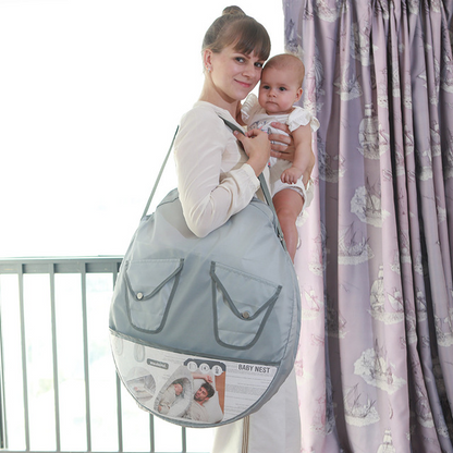 赤ちゃんを抱っこしたママが折りたたみ蚊帳付きベビーベッドを丸型バッグに入れて肩掛けしている写真（ポータブルベビーベッドの持ち運びイメージ）
