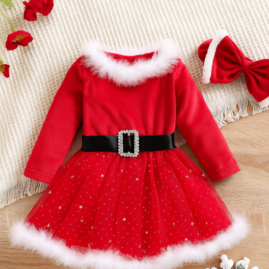 赤いベビークリスマスドレス。白いフェイクファーの襟と裾、真珠のバックル付き黒いベルト、金の星柄チュールスカートが特徴。木目調の背景に置かれ、周囲にクリスマスの装飾が添えられ、温かみのある祝祭的な雰囲気を演出している。
