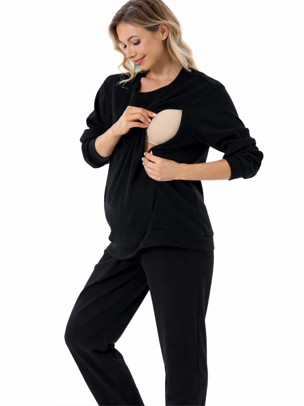 黒の授乳対応スウェットとパンツを着た女性が、胸元の授乳用開口部を手で開いて内側のベージュのインナーを見せている。白背景のスタジオ撮影。
