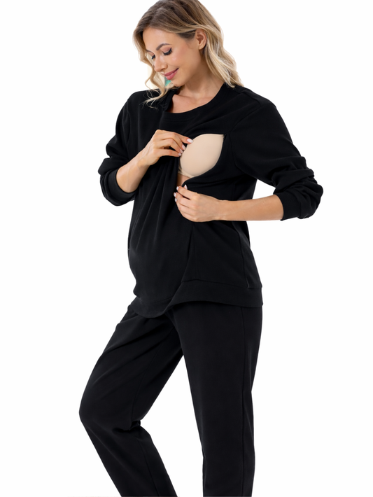 黒の授乳対応スウェットとパンツを着た女性が、胸元の授乳用開口部を手で開いて内側のベージュのインナーを見せている。白背景のスタジオ撮影。
