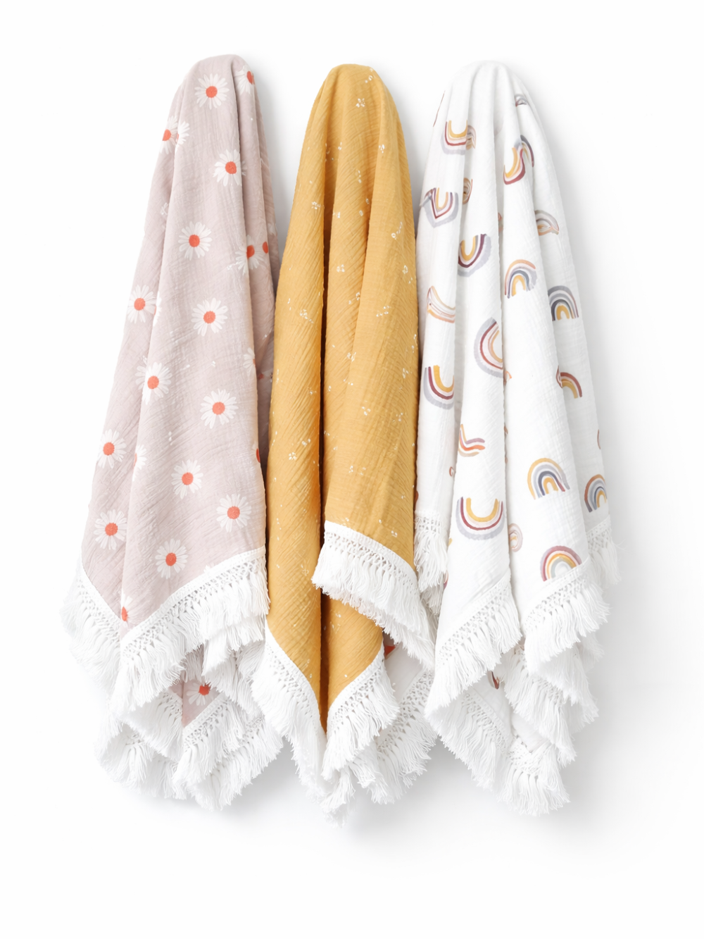 白背景に、フリンジとレース縁取りのモスリンブランケット3枚（デイジー柄のくすみピンク、花柄のマスタード、レインボー柄のホワイト）が並んで吊り下げられた商品画像。
