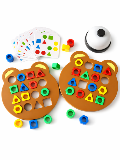 白背景のスタジオ撮影。クマ型の型はめボードが2枚並び、赤・青・黄・緑のカラフルな形パーツが差し込まれている。左上にお題カードの束、右上に白いベルがあり、周囲に追加のパーツが散らばっている。
