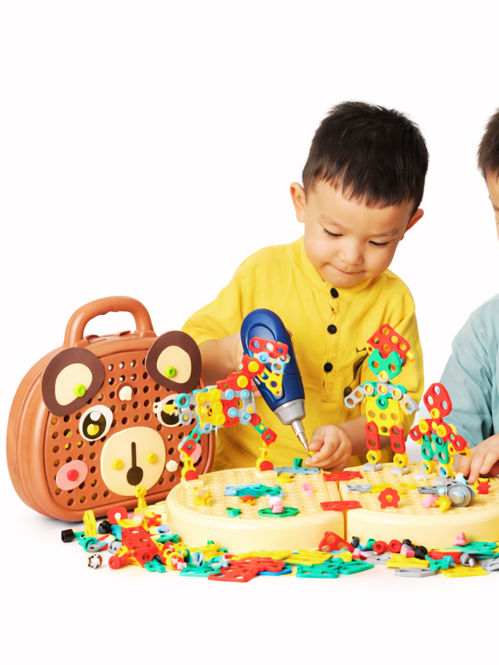 白背景のスタジオ風イメージ。ドリル付きの組み立て玩具セットで遊ぶ子どもが写っており、テーブル上にカラフルなパーツ、円形の土台、組み立てたロボット状の作品、左側にクマ型の収納ケースが配置されている。
