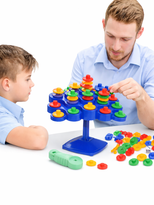 白背景の撮影イメージ。バランスゲームのおもちゃ（青い台座とボード）に、赤・黄・緑のコマを積み上げて遊んでいる様子で、周囲に予備のコマやサイコロ、パーツが置かれている。
