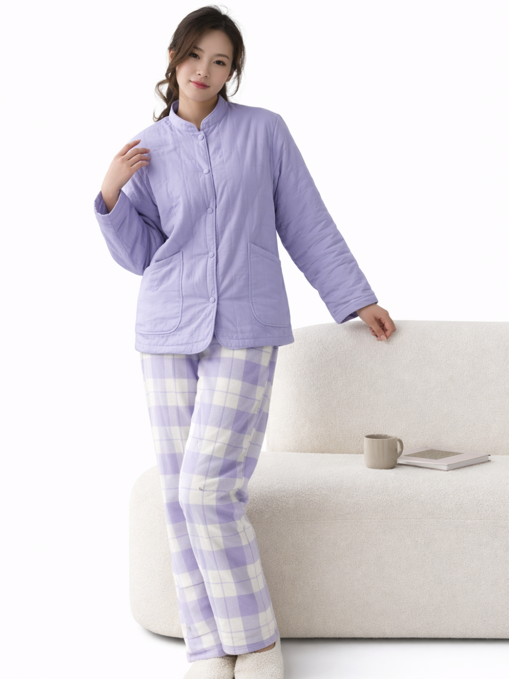 白背景のスタジオで撮影された、ラベンダー色の中綿入りルームウェア上着とチェック柄パンツを着た女性の着用イメージ。ソファにもたれて立っている。
