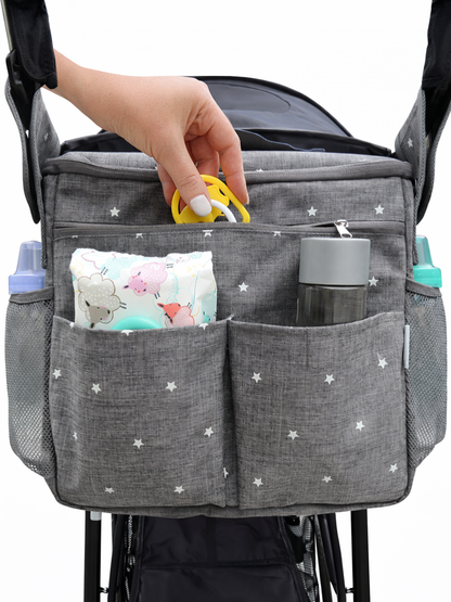白背景のスタジオで撮影された、星柄のグレーのベビーカー用オーガナイザー。複数ポケットにおしりふきとボトルを収納し、手が黄色い小物を上部ポケットに入れている。
