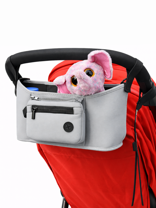 白背景のスタジオで撮影された、赤いベビーカーに取り付けたライトグレーのベビーカー用オーガナイザー。前面にファスナーポケットがあり、内側からピンクのぬいぐるみがのぞいている。
