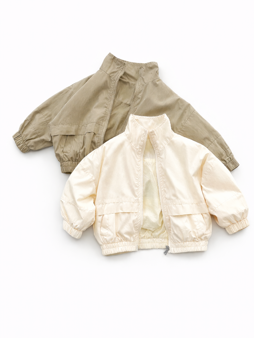 白背景で、キッズ用の軽量ジップジャケット2色（カーキ／アイボリー）を平置きで斜めに重ねて撮影した商品画像。スタンドカラーとゴム入り袖口・裾のデザインが分かる。