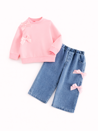 白背景で、女の子用2点セット（ピンクのリボン付きスウェット＋ライトブルーのデニムパンツ）を平置きで撮影した商品画像。トップスは肩と胸元にリボン装飾、パンツは脚にリボンが付いたデザイン。