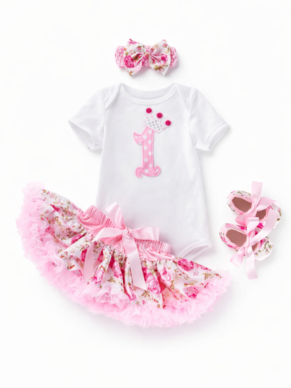 白背景に、ピンクの「1」と王冠アップリケ入り白ロンパース、ピンクの花柄チュチュスカート（サテンリボン付き）、花柄リボンのヘアバンド、ピンクリボン付き花柄ベビーシューズが配置された商品画像。
