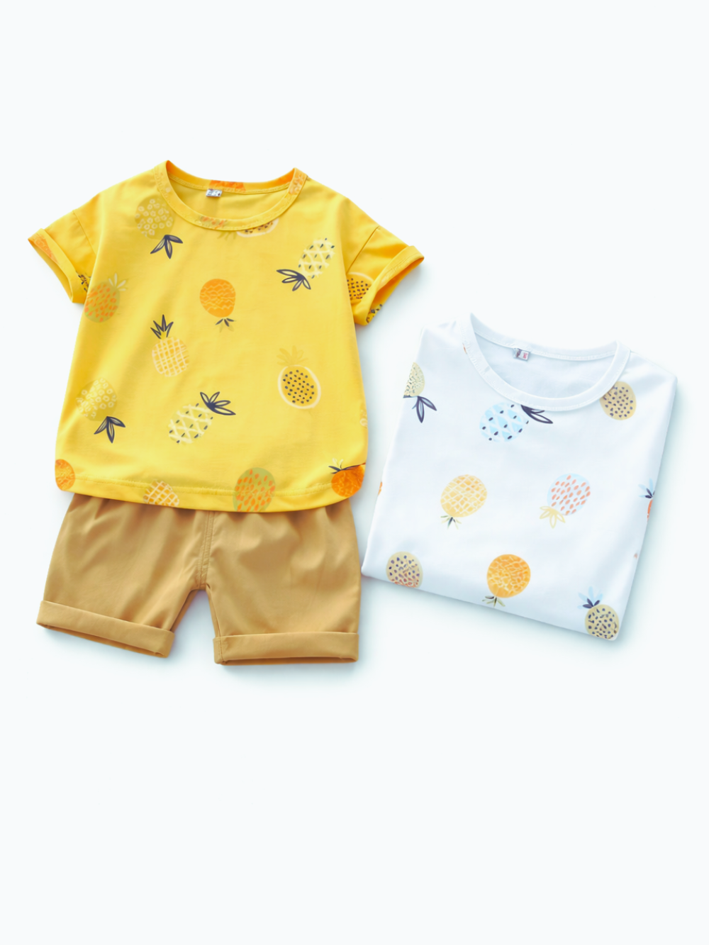 白背景に、パイナップル柄の子ども用夏服セットが配置された商品画像。イエローのパイナップル柄半袖Tシャツとベージュのショートパンツ、右側に同柄のホワイト半袖Tシャツが折りたたまれて置かれている。
