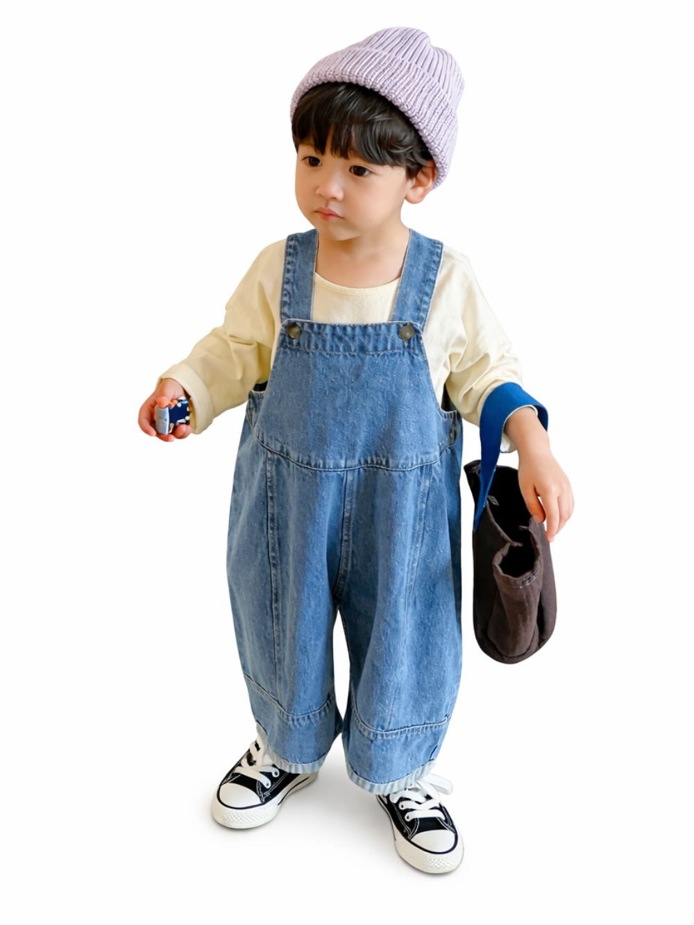 白背景で、ラベンダー色のニット帽をかぶった幼児が、クリーム色の長袖トップスとライトブルーのデニムオーバーオール、黒白スニーカーを着用し、手に小さなおもちゃとブラウンのバッグを持って立っている商品写真。

