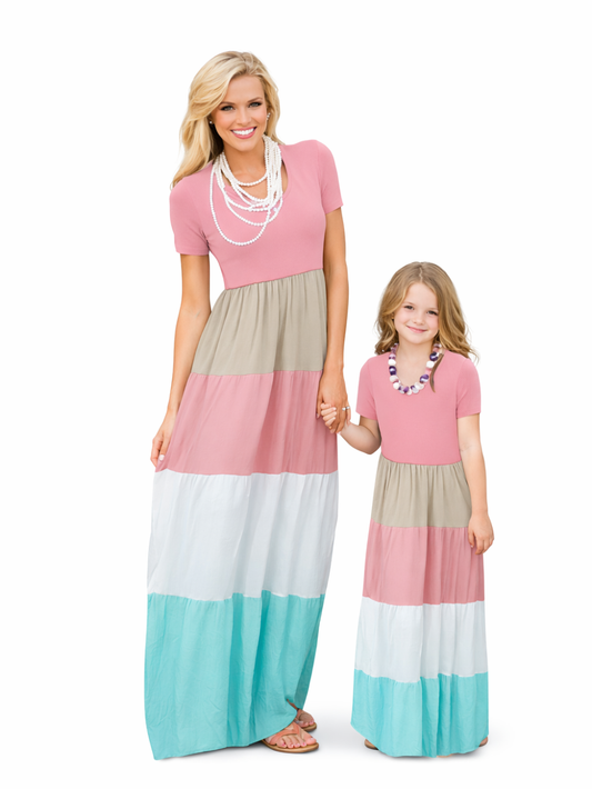 白い背景の前で手をつないで立つ母娘のリンクコーデ。ピンク×ベージュ×ホワイト×ターコイズの配色切替マキシワンピースを着用し、母は白いパール風ネックレスとサンダル、娘はビーズ風ネックレスを合わせている。
