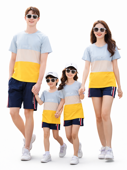 家族4人（両親と子ども2人）が、おそろいの水色×ベージュ×イエローのカラーブロックTシャツとネイビーのショートパンツ、白い帽子とサングラスを身に着け、白背景のスタジオで手をつないで並んでいる。
