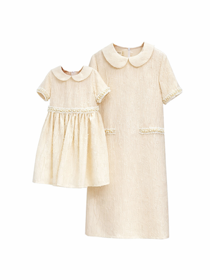 クリーム色のレース素材の親子おそろいワンピース（大人用1枚＋子ども用1枚）が、純白の背景に並んで写っている商品画像。丸襟と半袖、袖口・ウエスト・ポケット部分にパール風トリムが施されている。

