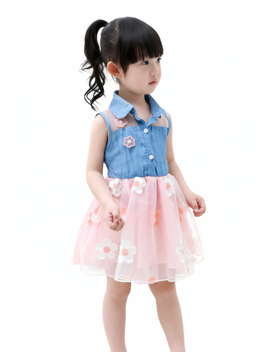 白い無地背景のスタジオ撮影で、ポニーテールの幼い女の子が、ブルーのノースリーブデニムトップスと花モチーフ付きのピンクのチュールスカートを着用して立っている。
