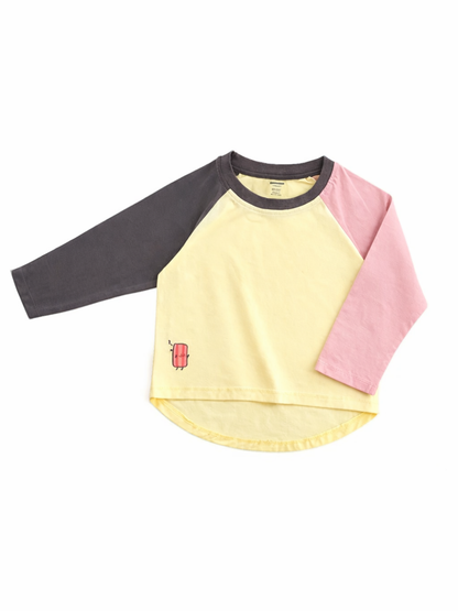 淡いイエローの身頃に、左袖はチャコールグレー、右袖はピンクのラグラン長袖キッズTシャツ。裾付近に赤いアイスキャンディの小さなワンポイントがあり、白背景で平置き撮影されている。
