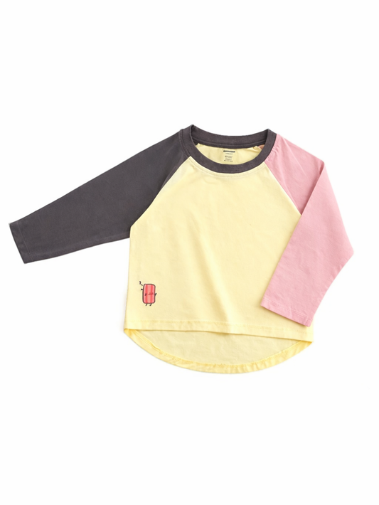 淡いイエローの身頃に、左袖はチャコールグレー、右袖はピンクのラグラン長袖キッズTシャツ。裾付近に赤いアイスキャンディの小さなワンポイントがあり、白背景で平置き撮影されている。
