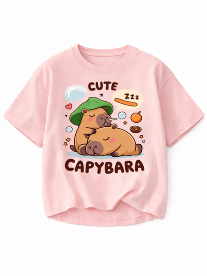 淡いピンクの半袖Tシャツ（クルーネック）。前面に、眠るカピバラ2匹のイラストと「CUTE CAPYBARA」の文字、ハートやフルーツなどの小さなモチーフがプリントされたデザイン。背景は白。

