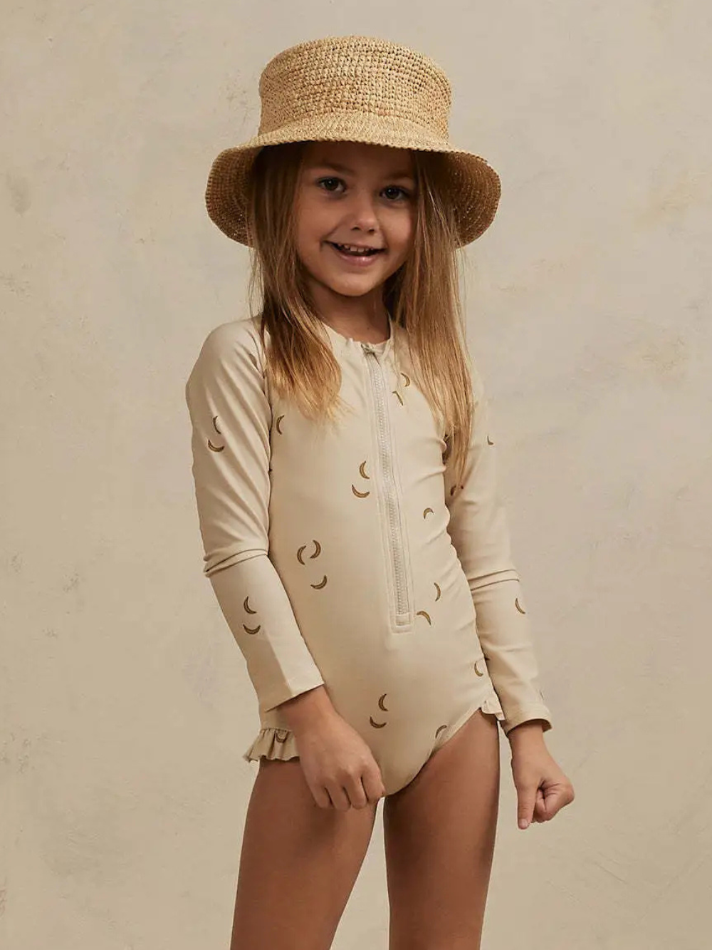 麦わら帽子をかぶった女の子が、白地に三日月柄の長袖ラッシュガード一体型水着（フロントジップ、裾フリル付き）を着て、白い背景の前で立っている。
