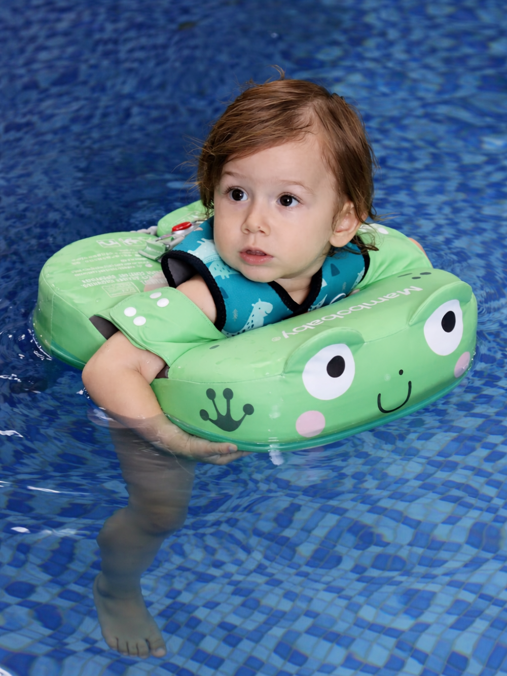 幼児がカエルの顔デザインのベビー用浮き輪に入り、青いプールの水面に浮かんでいる様子。
