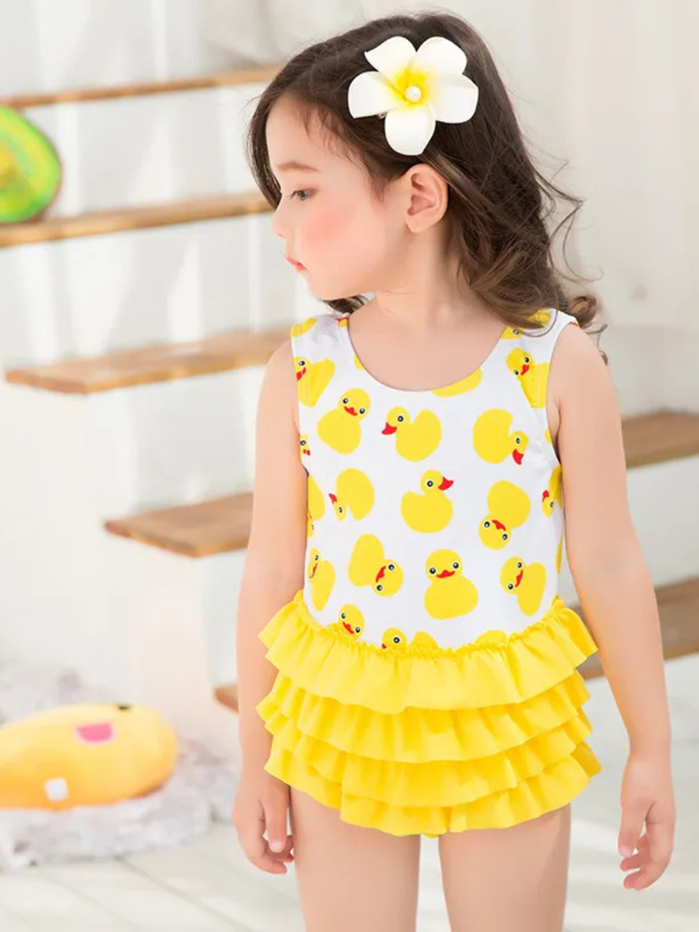 女の子がアヒル柄の子ども用ワンピース水着を着用している。白地に黄色いアヒルのプリント、ウエスト部分は黄色の3段フリルスカート風デザイン。室内の明るい背景で、木の階段と小物が写っている。