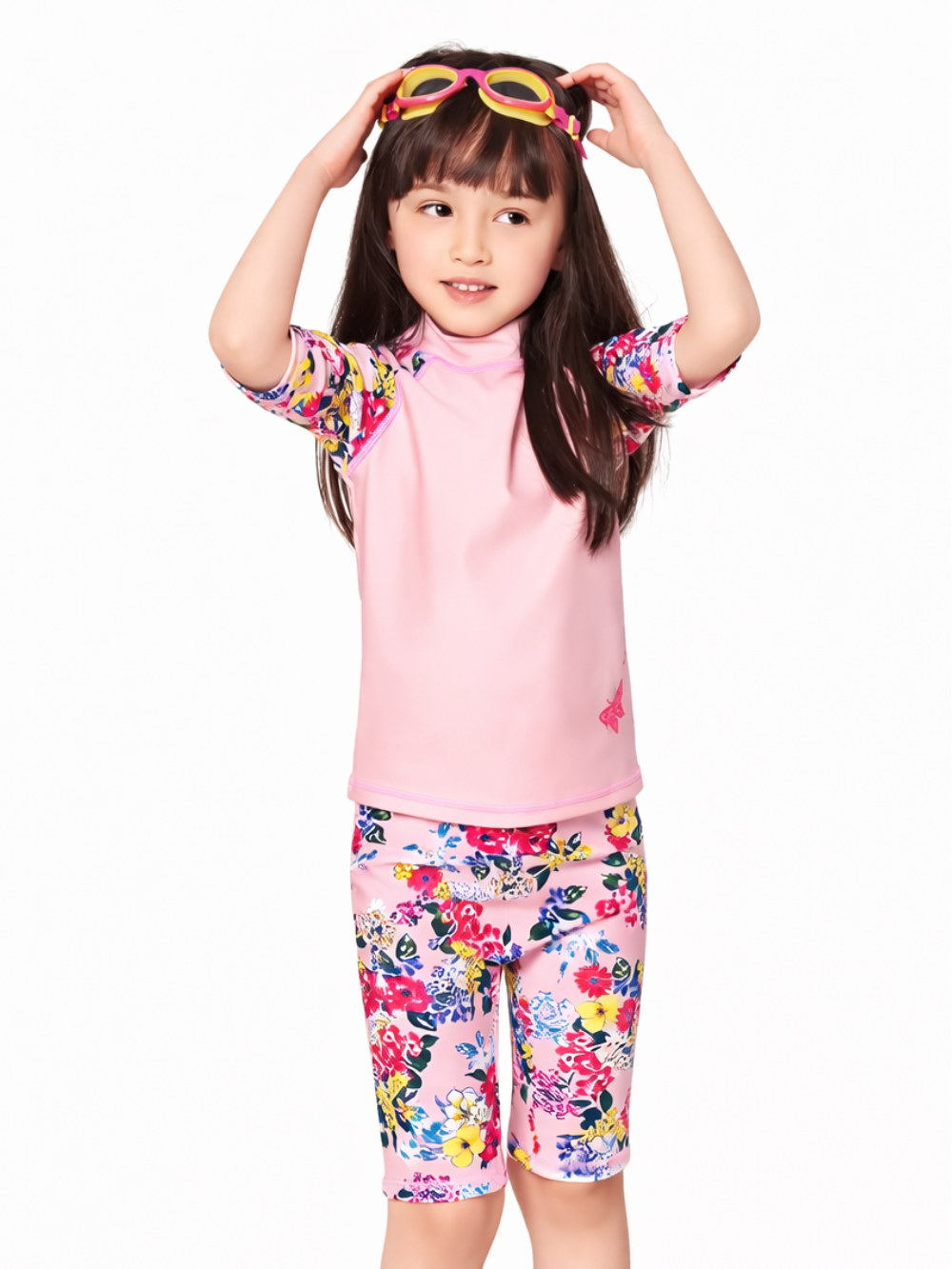 白背景のスタジオ撮影で、女の子がピンクのラッシュガード上下セット水着を着用している。トップスはピンク無地で袖が花柄、ボトムは花柄の膝丈レギンス風。頭に黄色×ピンクのゴーグルをのせ、両手で調整しているポーズ。
