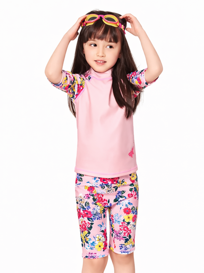 白背景のスタジオ撮影で、女の子がピンクのラッシュガード上下セット水着を着用している。トップスはピンク無地で袖が花柄、ボトムは花柄の膝丈レギンス風。頭に黄色×ピンクのゴーグルをのせ、両手で調整しているポーズ。