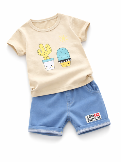白背景のスタジオ撮影で、ベビー／キッズ用の上下2点セットをフラットに配置した商品画像。アイボリーの半袖Tシャツにサボテンのイラストプリント、ボトムはブルーのショートパンツ（ウエストゴム、裾ロール風）。自然な薄い影付き。
