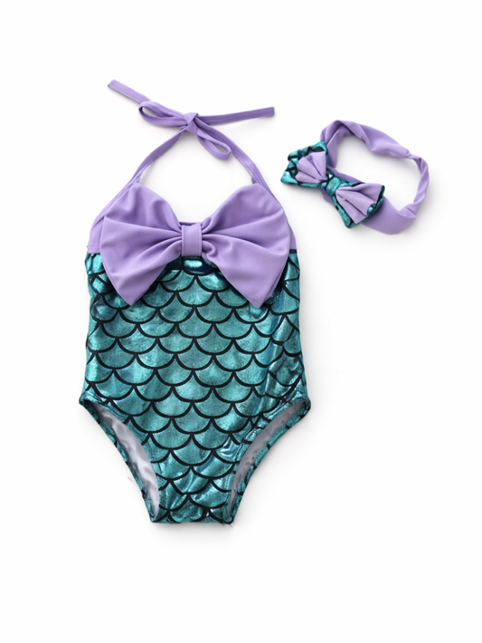 キラキラしたティールブルーのうろこ柄ワンピース水着と、同柄のリボン付きヘアバンドのセット。ラベンダーのホルターネック紐と大きなフロントリボンが特徴で、白背景に商品を並べたカタログ写真。