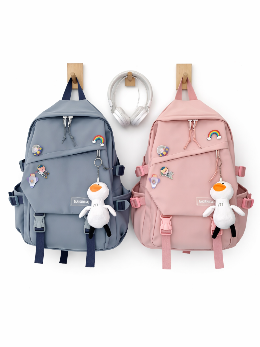 白背景に、グレーがかったブルーと淡いピンクのリュックが2点、正面向きで並んでいる。両方ともレインボーなどのピンバッジと、白いアヒルのぬいぐるみチャームが付いており、上部にフックで掛けられている。下に柔らかな影が入っている。
