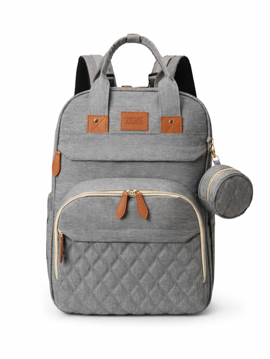 グレーのキルティング入りおむつバッグ（マザーズリュック）が、白い無地背景のスタジオ撮影で正面から写っている。ベージュのファスナーとブラウンのレザープル、前面ポケット、「SUNSHINE」ロゴ、サイドに吊り下げた丸型ボトルケースが見える。
