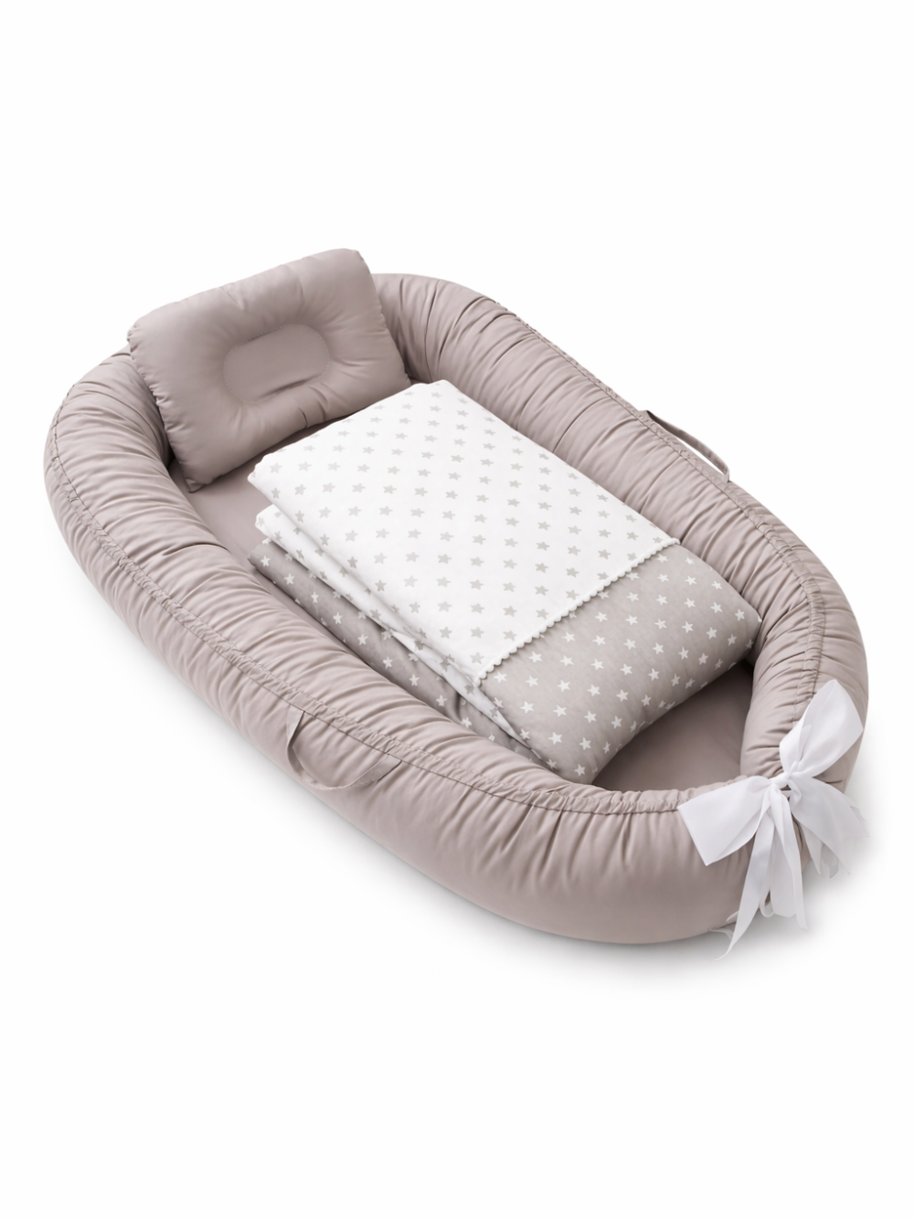 白い背景の上に、グレージュのベビーネスト（ベビー用クッションベッド）が斜め上から写っている。枕と星柄のブランケットが中に置かれ、端は白いリボンで結ばれている。
