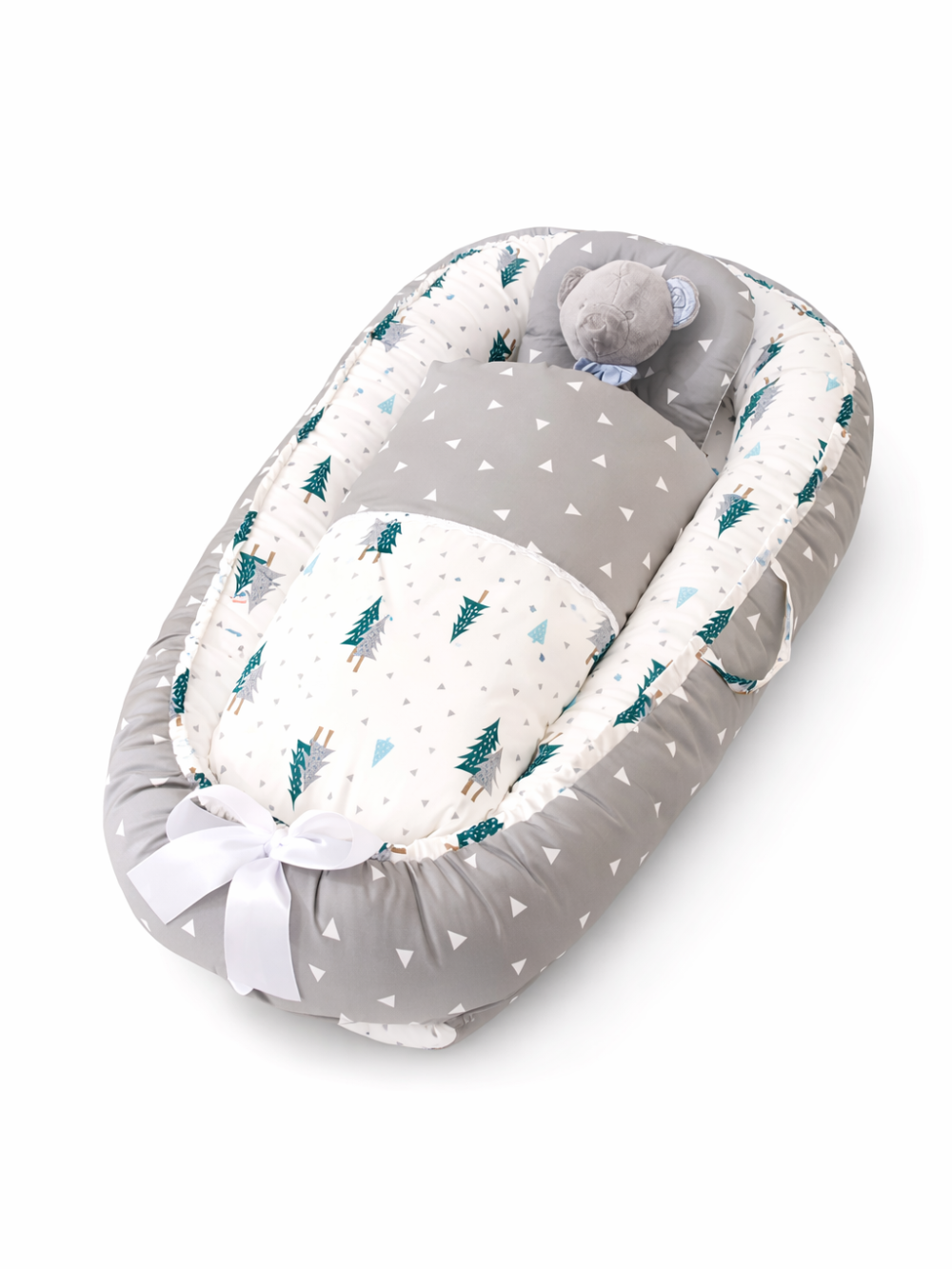 白い背景の上に、グレーと白のベビーネスト（ベビー用クッションベッド）が斜め上から写っている。三角柄と木のプリントが入り、足元は白いリボンで結ばれ、頭側にはクマのぬいぐるみが置かれている。
