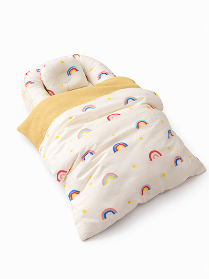 白い背景の上に、虹と星柄のクリーム色のベビーネスト兼ベビー布団セットが斜め上から写っている。中に同柄の枕があり、黄色いブランケットと掛け布団が重ねられている。