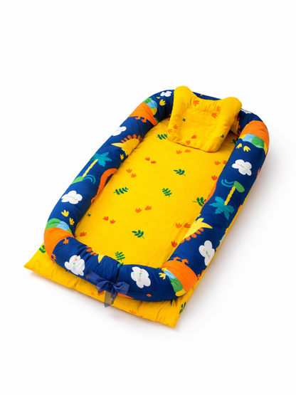 白い背景の上に、黄色のベビーネスト（ベビー用クッションベッド）が斜め上から写っている。内側は小花と葉の柄、外側はネイビーに雲や恐竜などのカラフルな柄が入り、枕付きで足元にリボンが結ばれている。