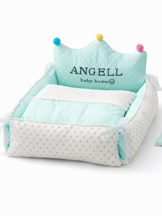 白い背景の上に、上から見たミントグリーンのベビーベッド（クラウン形ヘッドボード付き）が写っている。白×グレーの星柄サイドと水玉の掛け布団がセットになっており、ヘッドボードにはカラフルなポンポンが3つ付いている。横に同色の小さなクッションが置かれている。
