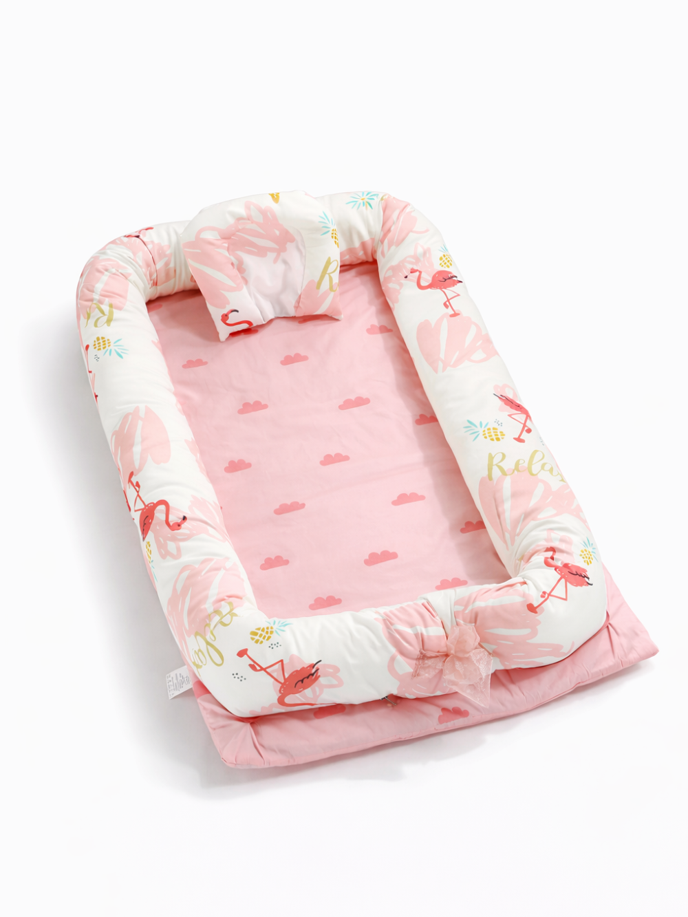 白い背景の上に、斜め上から見たピンクのベビーネスト（ベビー用クッションベッド）が置かれている。白地のサイドクッションにはフラミンゴとパイナップルの柄が入り、内側は雲柄のピンクマットで、枕とリボンが付いている。
