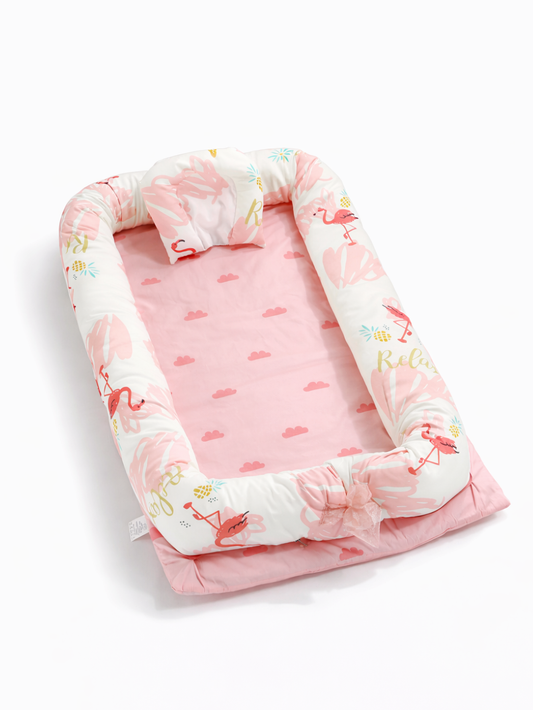 白い背景の上に、斜め上から見たピンクのベビーネスト（ベビー用クッションベッド）が置かれている。白地のサイドクッションにはフラミンゴとパイナップルの柄が入り、内側は雲柄のピンクマットで、枕とリボンが付いている。
